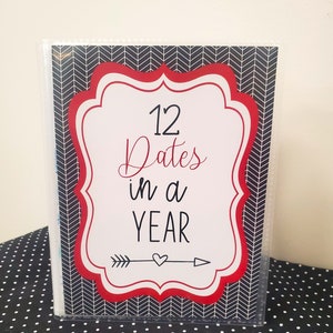 May include: A clear plastic binder with a cover featuring the text "12 Dates in a YEAR." The cover has a white and red decorative border over a black and white chevron pattern. An arrow and heart graphic are below the text.