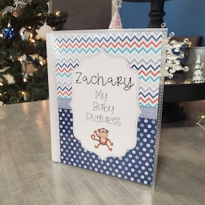 May include: A blue and white photo album with a chevron and polka dot pattern. The album has a white label with the text "Zachary My Baby Pictures" and a cartoon monkey.