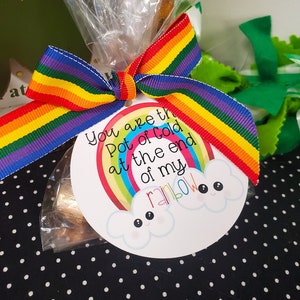 Puede incluir: Una bolsa de regalo atada con una cinta de arcoíris y una etiqueta redonda. La etiqueta presenta un arcoíris y dos nubes sonrientes con el texto "You are the Pot of gold at the end of my rainbow."