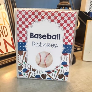 Peut inclure: Un album photo blanc avec un design sur le th&egrave;me du baseball rouge, blanc et bleu. La couverture de l'album pr&eacute;sente un ballon de baseball, des battes, des gants et des &eacute;toiles. Le texte "Baseball Pictures" est imprim&eacute; sur la face avant de l'album.