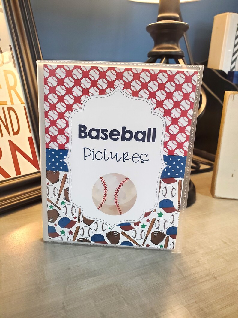 Peut inclure: Un album photo blanc avec un design sur le th&egrave;me du baseball rouge, blanc et bleu. La couverture de l'album pr&eacute;sente un ballon de baseball, des battes, des gants et des &eacute;toiles. Le texte "Baseball Pictures" est imprim&eacute; sur la face avant de l'album.