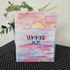 May include: A clear plastic photo album with a watercolor design. The cover features a sunset over the ocean with the words "SUMMER 2025". The sky is pink and the water is blue and pink. The album is on a black and white polka dot surface.