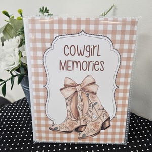 May include: A pink and white gingham scrapbook with the text "cowgirl memories" and a pair of brown boots with floral designs and a brown bow.