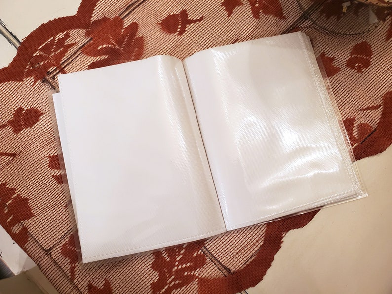 Peut inclure: Un album photo blanc avec des pochettes en plastique transparent pour ranger des photos. L'album est ouvert et pos&eacute; &agrave; plat sur une surface &agrave; motifs bruns.