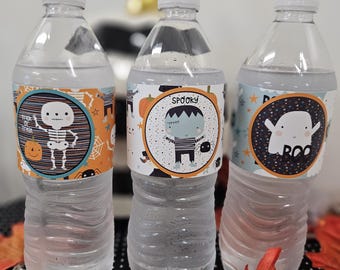 May include: Three clear water bottles with Halloween-themed labels. The labels feature a skeleton, a Frankenstein's monster, and a ghost with the word "BOO". The bottles are on a black and white polka dot surface with orange leaves.
