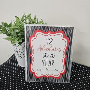 May include: A black and white chevron patterned card with a red border and a white heart. The card says "12 Adventures in a Year" with an arrow pointing to the right.