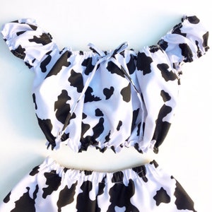Puede incluir: Un top corto de hombros descubiertos con estampado de vaca blanco y negro con un lazo en el escote y pantalones cortos a juego.