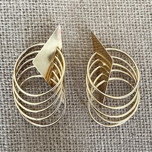 May include: A pair of gold-toned earrings featuring a flat, diamond-shaped metal plate attached to a series of four concentric circular wire hoops. The design is symmetrical, with the hoops suspended from the top point of the diamond-shaped base.