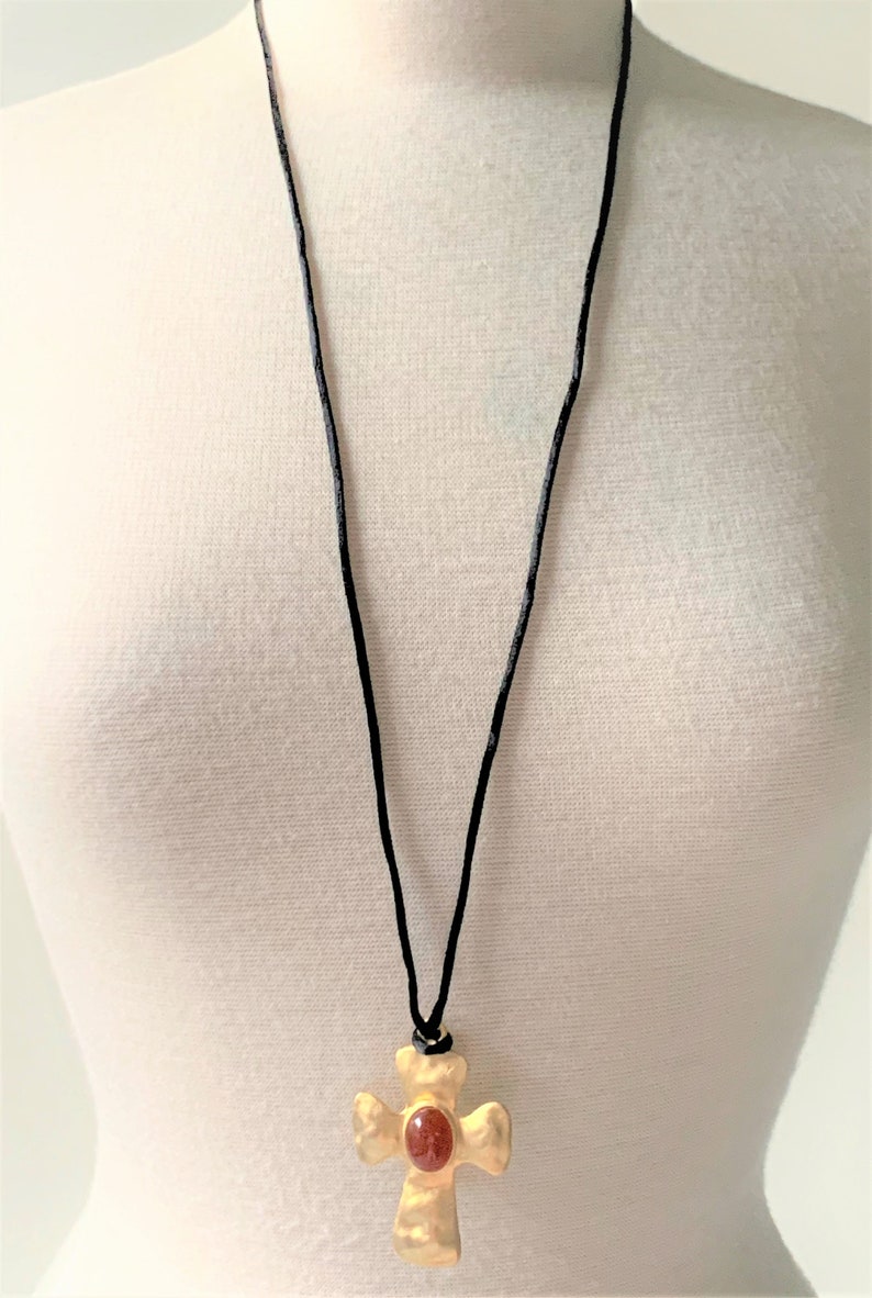 May include: A gold cross pendant necklace with a red gemstone in the center, hanging from a black cord.