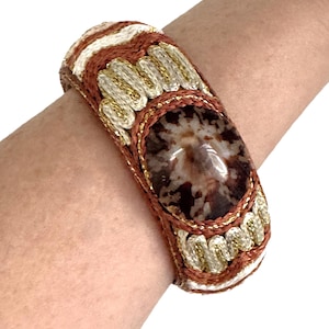 May include: A wide, handcrafted bracelet featuring a large, oval-shaped, speckled brown stone at its center. The bracelet is made with woven brown, white, and gold-colored threads, creating a textured and ornate design. The bracelet is worn on a wrist.