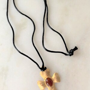 May include: A gold cross pendant with a red gemstone set in the center, hanging from a black cord necklace.