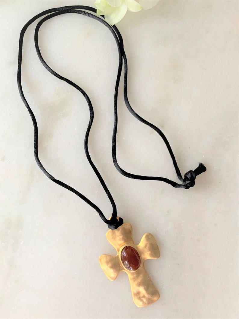 May include: A gold cross pendant with a red gemstone set in the center, hanging from a black cord necklace.