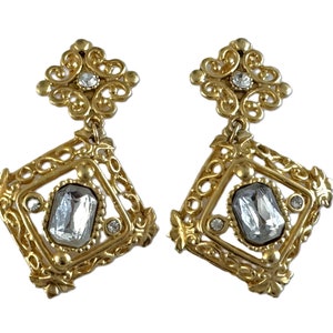 May include: Gold tone dangle earrings with a large clear faceted stone set in a square frame with ornate filigree details.