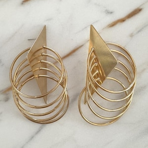May include: A pair of gold-toned earrings featuring a flat triangular metal plate attached to a series of four concentric circular wire hoops. The earrings have a post-back closure and exhibit a polished metallic finish with geometric shapes.