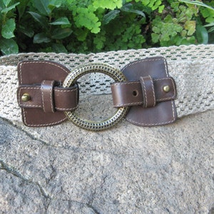 May include: A beige crochet belt with brown leather accents and a large, circular, brass-toned buckle. The belt features a woven design and leather details around the buckle and clasp. The belt is displayed on a stone surface.
