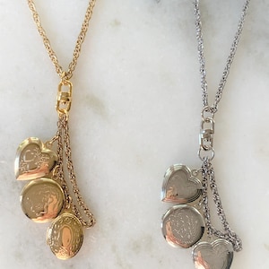 May include: Two necklaces with heart and oval-shaped lockets. One necklace is gold-toned, the other is silver-toned. Each necklace has a chain with a clasp and a cluster of three lockets with intricate designs.