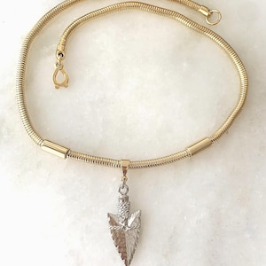 May include: Gold-toned necklace with a snake chain design. The necklace features a pendant shaped like an arrowhead with a textured silver finish. The necklace has a lobster clasp closure.