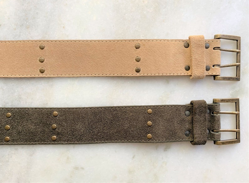 May include: Two suede belts with brass buckles and studs. The top belt is light brown and the bottom belt is dark green.