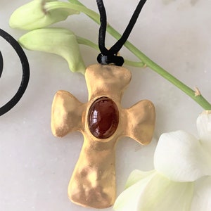 May include: A gold cross pendant with a red gemstone set in the center, hanging from a black cord.
