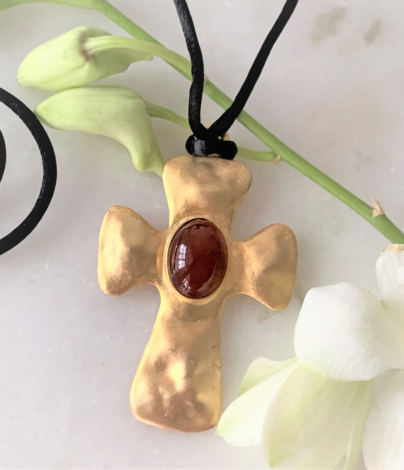 May include: A gold cross pendant with a red gemstone set in the center, hanging from a black cord.