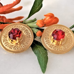 May include: A pair of gold earrings with a red gemstone in the center of each. The earrings have a detailed design around the gemstone.