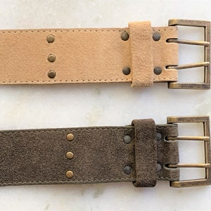May include: Two suede belts with brass buckles and decorative studs. The top belt is light brown and the bottom belt is dark brown.