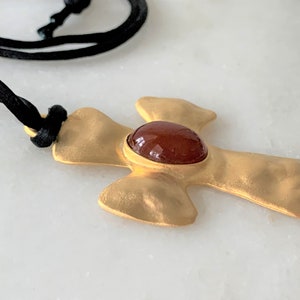 May include: A gold-toned cross pendant with a red gemstone in the center, hanging from a black cord.