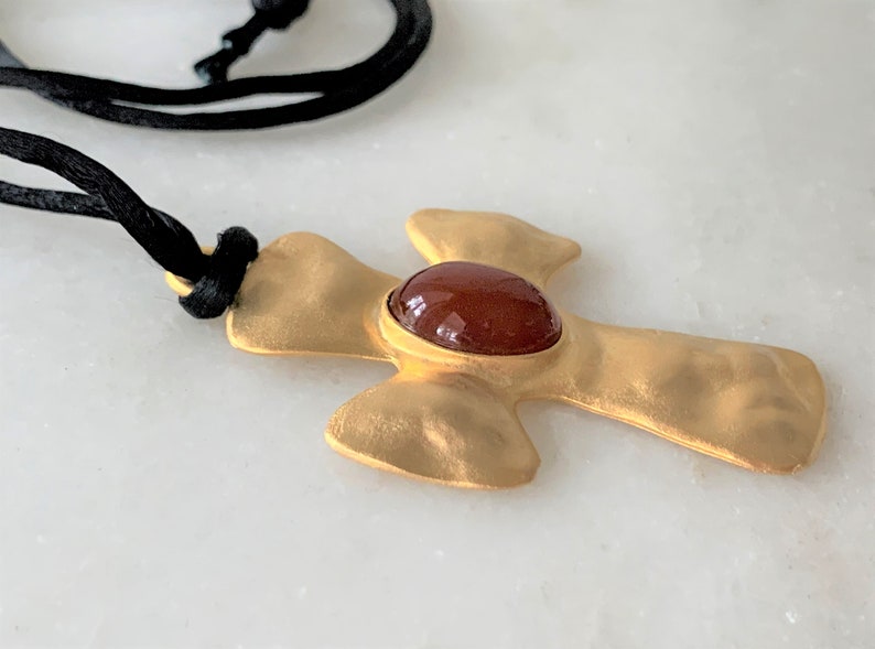May include: A gold-toned cross pendant with a red gemstone in the center, hanging from a black cord.