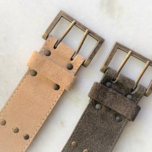 May include: Two suede belts with brass buckles. One belt is light brown and the other is dark brown.
