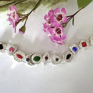 May include: A silver-toned bracelet with various colorful gemstones, including red, green, blue, and black. The bracelet features a decorative design with floral and geometric shapes. Pink flowers and greenery are in the background.