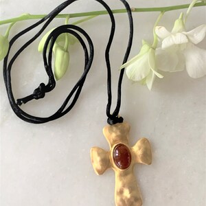 May include: A gold cross pendant with a red gemstone in the center, hanging from a black cord.