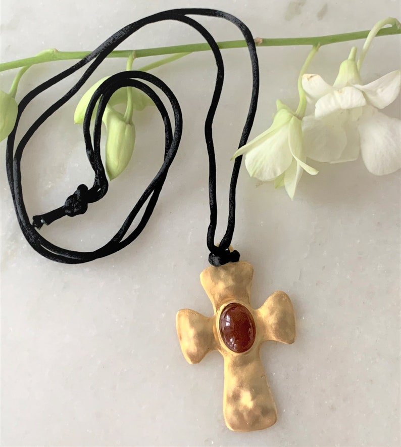 May include: A gold cross pendant with a red gemstone in the center, hanging from a black cord.