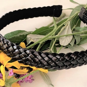 Peut inclure: Un serre-t&ecirc;te tress&eacute; noir en cuir. Le serre-t&ecirc;te est pos&eacute; sur un lit de feuilles vertes et de fleurs jaunes.