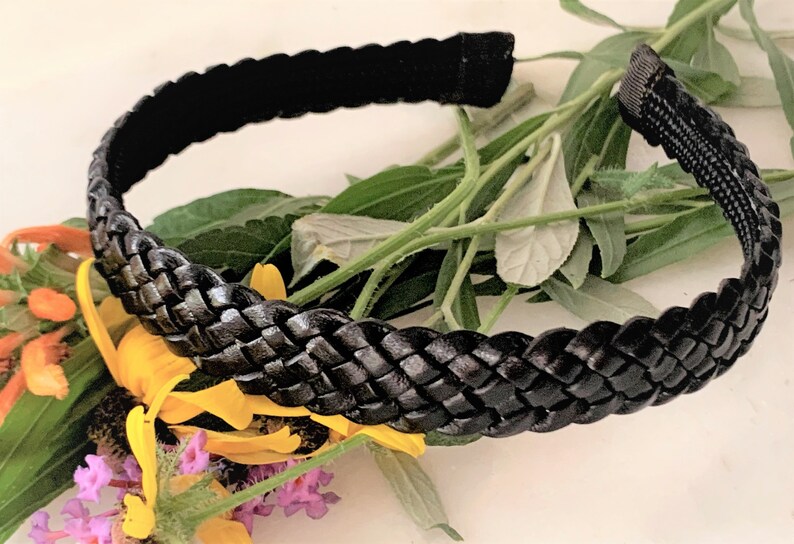 Peut inclure: Un serre-t&ecirc;te tress&eacute; noir en cuir. Le serre-t&ecirc;te est pos&eacute; sur un lit de feuilles vertes et de fleurs jaunes.
