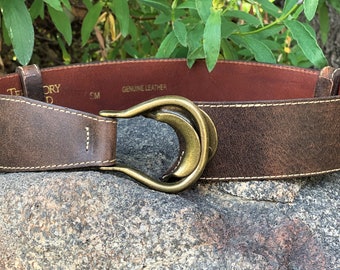May include: A brown leather belt with a gold-toned horseshoe buckle. The belt is stitched with a light brown thread and has the text "Genuine Leather" printed on the inside.