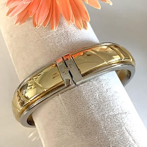 May include: A gold and silver bangle bracelet with a textured design. The bracelet is open and has a hinged clasp.