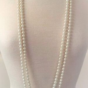 May include: A long, white pearl necklace with three strands. The necklace is draped over a white mannequin.