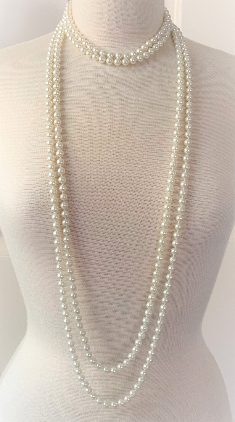 May include: A long, white pearl necklace with three strands. The necklace is draped over a white mannequin.