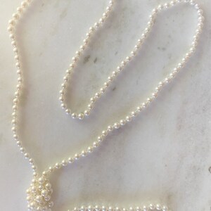May include: A long strand of white pearl beads, knotted together, laid out on a white marble surface.