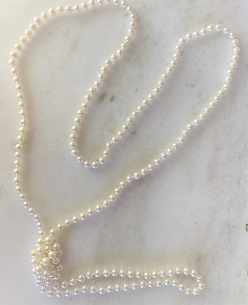 May include: A long strand of white pearl beads, knotted together, laid out on a white marble surface.
