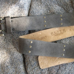 May include: A gray suede belt with gold-toned studs and a double-prong buckle. The belt is labeled "ELITE ACCESSORIES" and "5717".