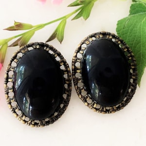 May include: A pair of vintage oval-shaped clip-on earrings. Each earring features a glossy black center stone set within a decorative, openwork bronze-toned metal frame. The earrings are displayed on a white surface with floral accents.