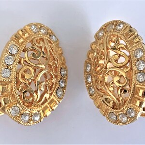 May include: A pair of gold-toned clip-on earrings. Each earring has an oval shape with a filigree design and is embellished with clear, round rhinestones. The earrings have a vintage aesthetic.