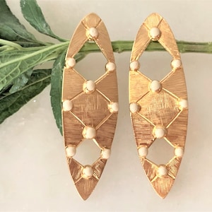 May include: A pair of gold-tone earrings with a geometric design and white accents. The earrings are oval-shaped and have a textured surface.