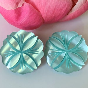 May include: Two light blue, flower-shaped cabochons with carved details. The cabochons have a scalloped edge and a central design of curved lines and pointed elements. A pink flower petal is in the background.