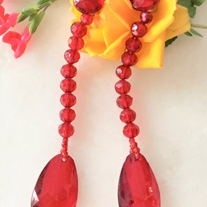 May include: A pair of long, red crystal earrings with teardrop-shaped drops. The earrings are made of faceted beads and have a round, red crystal at the top.