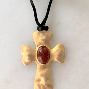 May include: A gold cross pendant with a red gemstone set in the center, hanging from a black cord.