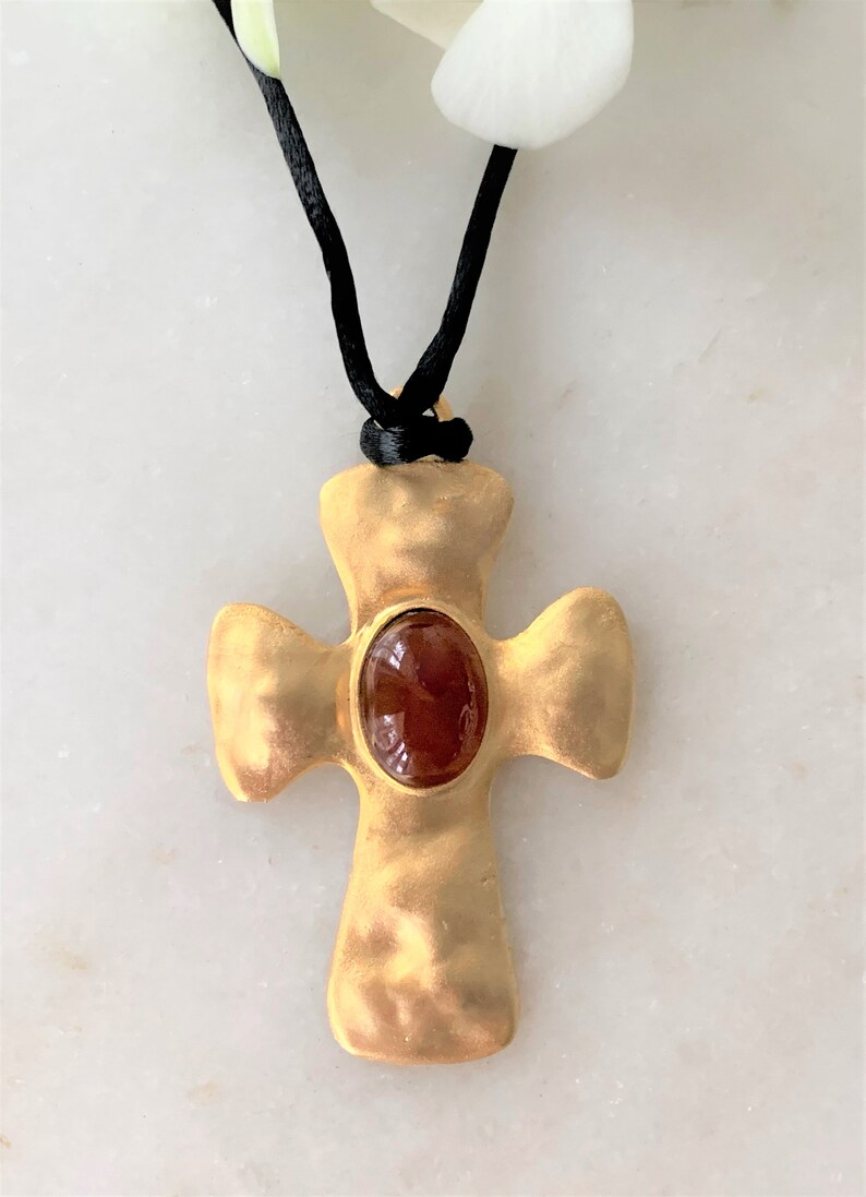 May include: A gold cross pendant with a red gemstone set in the center, hanging from a black cord.