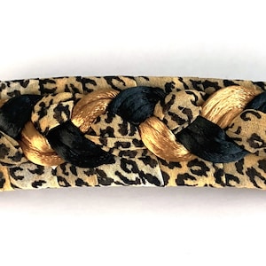 May include: A rectangular hair barrette with a leopard print fabric base. Braided black and gold ribbons are woven across the top. The barrette is approximately 5 inches long.