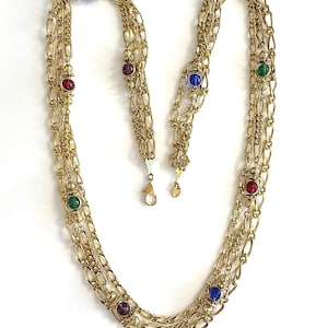 May include: Gold-toned necklace with three strands of linked chain. The necklace is adorned with colorful, round gemstones in red, blue, green, and purple. The necklace has a delicate, vintage aesthetic.